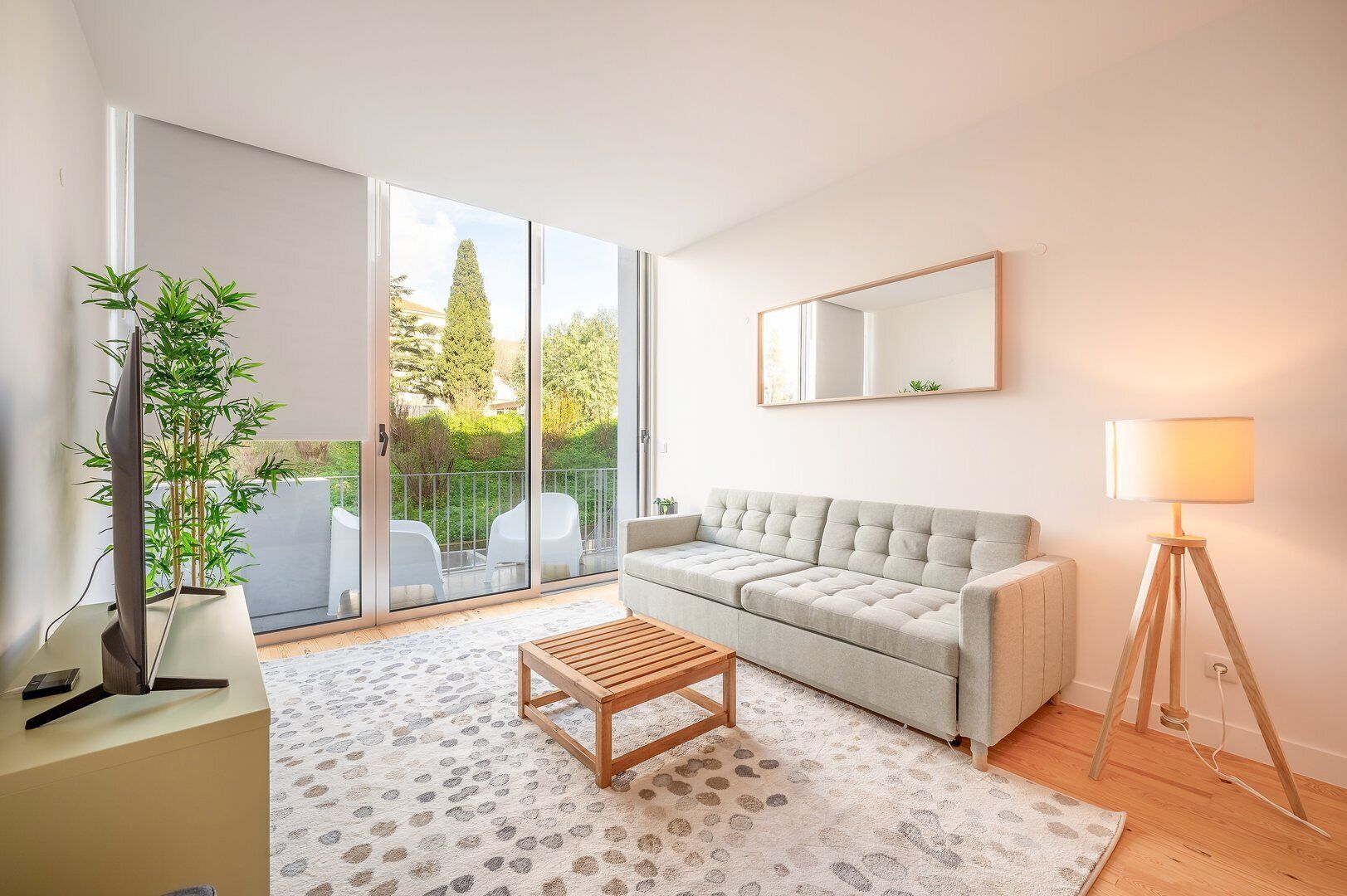 Een moderne woonkamer met een grijze bank, een houten salontafel, een vloerlamp en een grote schuifpui die leidt naar een balkon met tuinmeubels.