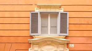 A building facade with a large wooden double door, a small plaque with the number 122, and two windows with shutters labeled "FAUSTO" and "SANTINI". - Frattina Atmosphere apartment (Rome)