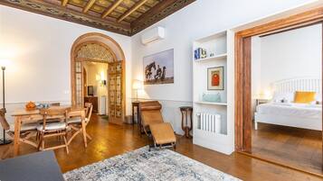 Ein Zimmer mit Holztisch, einer Couch, einem Sessel, einem Heizkörper, einem Bett und einer Decke mit aufwendigen Verzierungen.