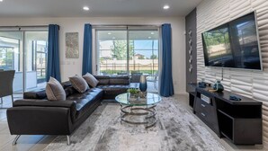 A modern living room with a sectional sofa, a glass coffee table, a TV on a wall, and large windows with curtains. - Beautiful and Cozy Home at Sonoma Resort (Kissimmee)