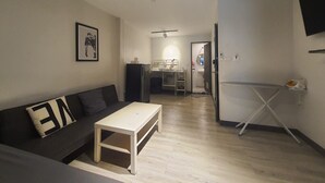 A compact living space with a sofa, a small table, a kitchenette, and a foldable ironing board.