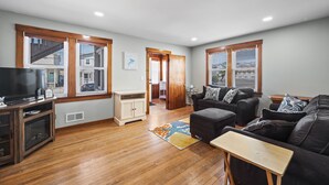 A spacious living room with hardwood floors, a large flat-screen TV, a fireplace, and a cozy seating area with a couch and ottoman.