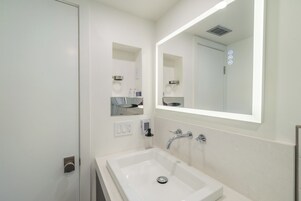 A modern bathroom with a white sink, a large mirror, and a wall-mounted soap dispenser.