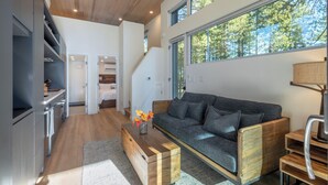 Ein modernes Wohnzimmer mit grauem Sofa, einem Holztisch und Blick auf Bäume durch große Fenster.
