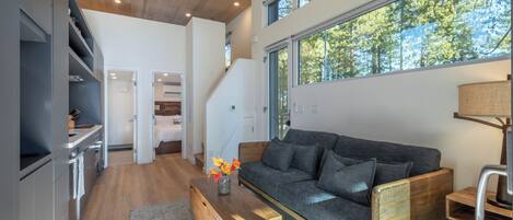 Un salón moderno con un sofá gris, una mesa de centro de madera y vistas a árboles a través de amplios ventanales.