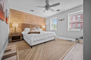 Une chambre avec un mur en briques, un lit avec une literie blanche, une table de chevet avec une lampe, un ventilateur de plafond et une fenêtre donnant sur un immeuble.