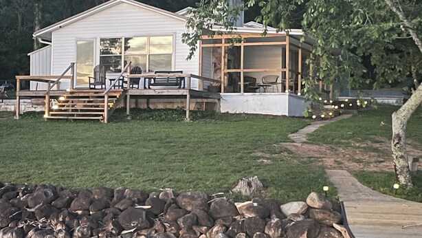Eine Blockhütte am Seeufer mit einer Terrasse, einer überdachten Sitzgelegenheit im Freien und einer Steinschutthalde.