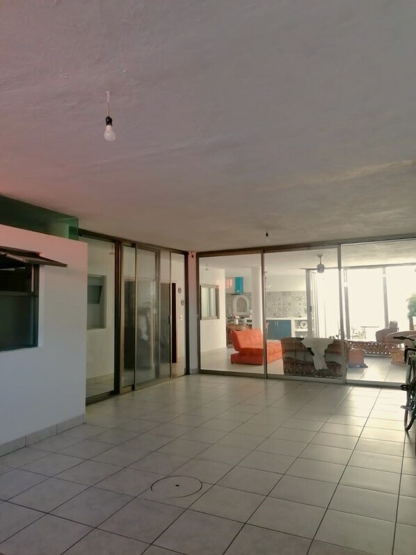 Amplia habitación con suelo de baldosas, puerta de cristal corredera que da a una sala de estar con sofá y bicicleta, y una cocina con estufa y armarios.