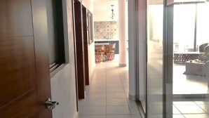 A hallway with a wooden door, a tiled floor, and a view into a room with a kitchen area.