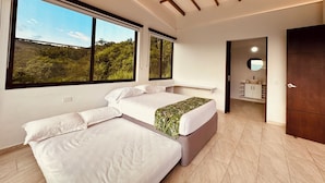 Une chambre moderne avec un grand lit, un lit plus petit et un canapé. De grandes fenêtres offrent une vue sur la verdure. La chambre est dotée d’un plafond en bois et d’un ventilateur de plafond.