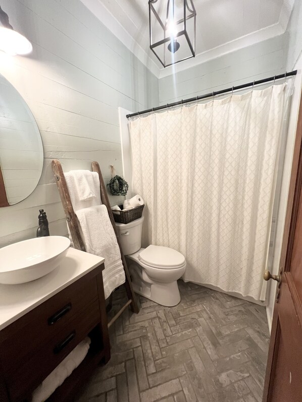 Une salle de bain avec un lavabo blanc, un meuble-lavabo en bois, des toilettes, un rideau de douche et un sol à motifs en chevrons.