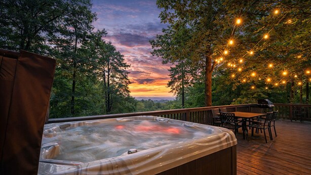 Een hottub op een houten terras met uitzicht op een zonsondergang, omgeven door bomen en lichtjes.