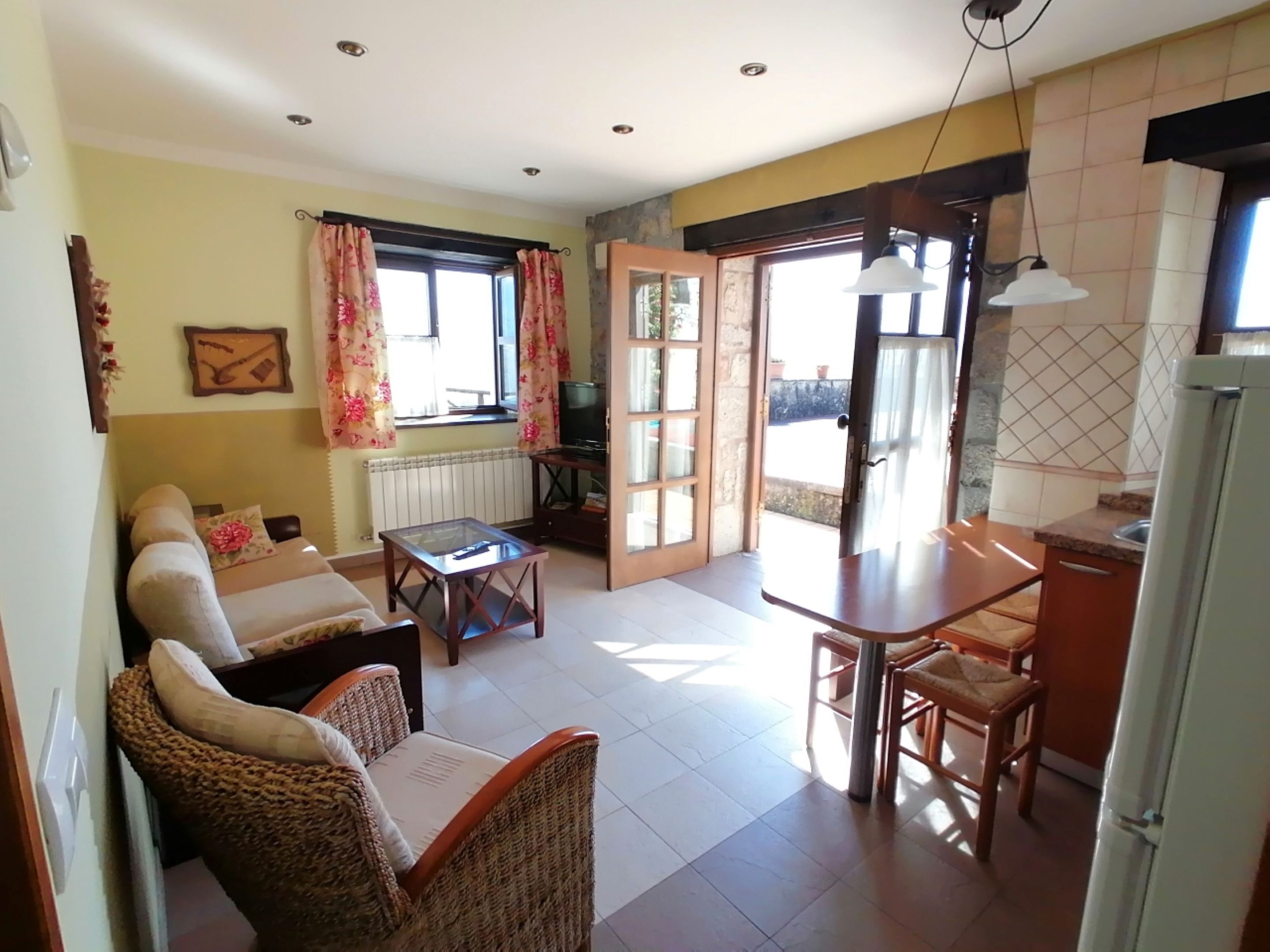 Una sala acogedora con sofá, una silla de mimbre, una mesa de centro de vidrio y un televisor. Hay una zona de comedor con una mesa y sillas, y una cocina con refrigerador y ventana.