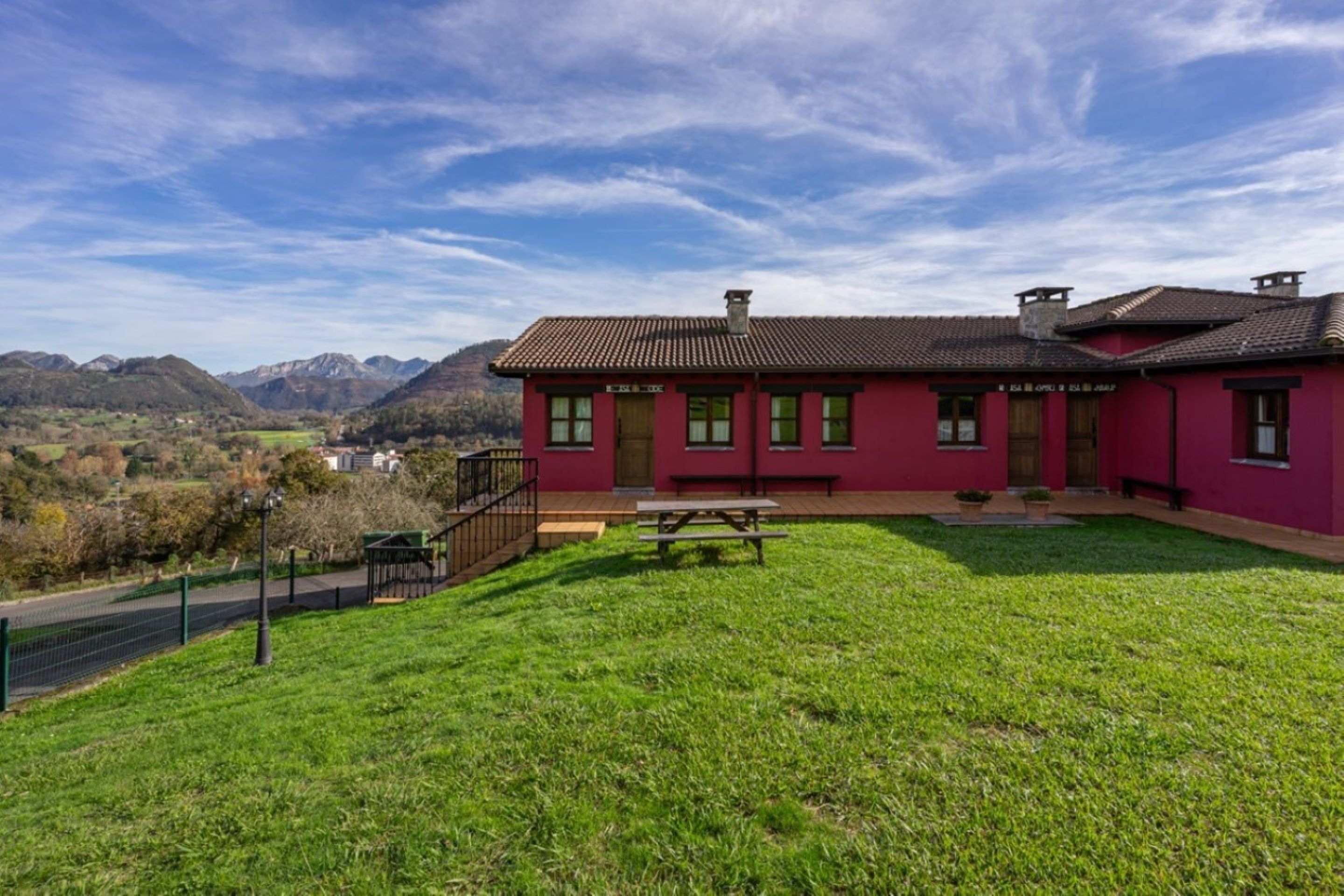A red building with a picnic table in the front yard, surrounded by a green lawn and mountains in the background.