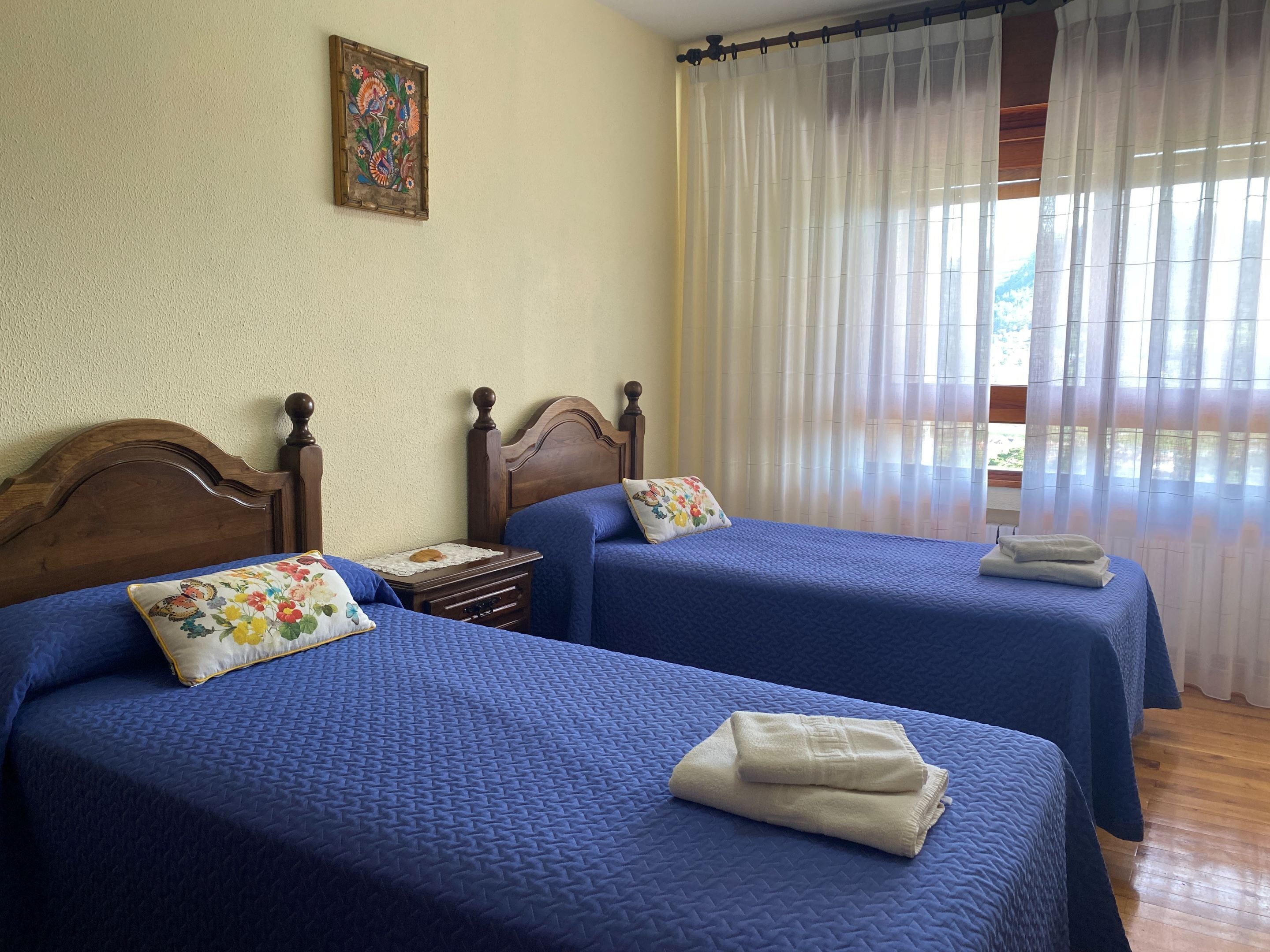 A room with two beds, each with a blue quilted bedspread and a floral pillow. There is a wooden headboard on each bed, and a small bedside table with a lamp. A painting hangs on the wall above the beds.