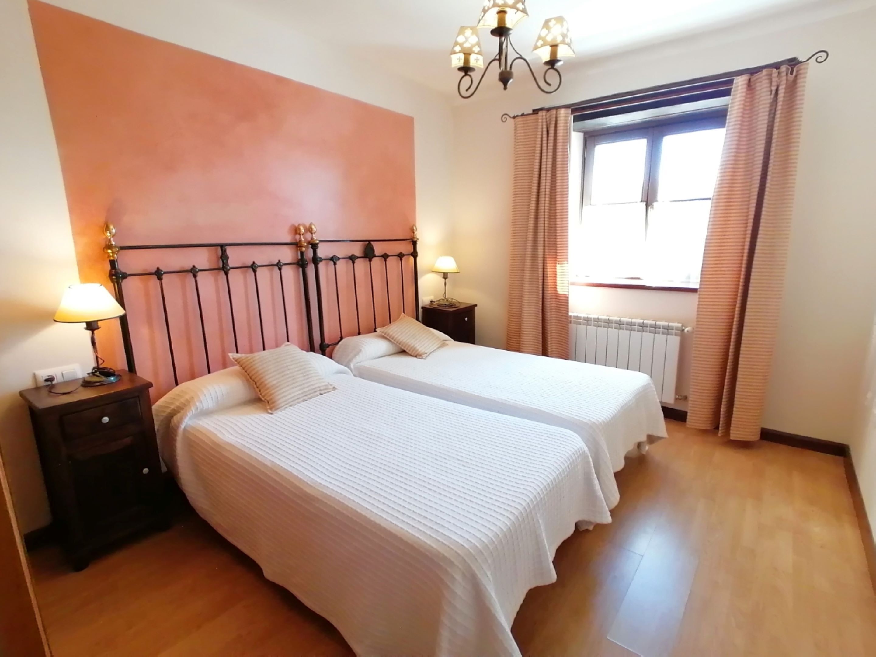 A bedroom with two beds, a chandelier, a window with curtains, and a radiator.