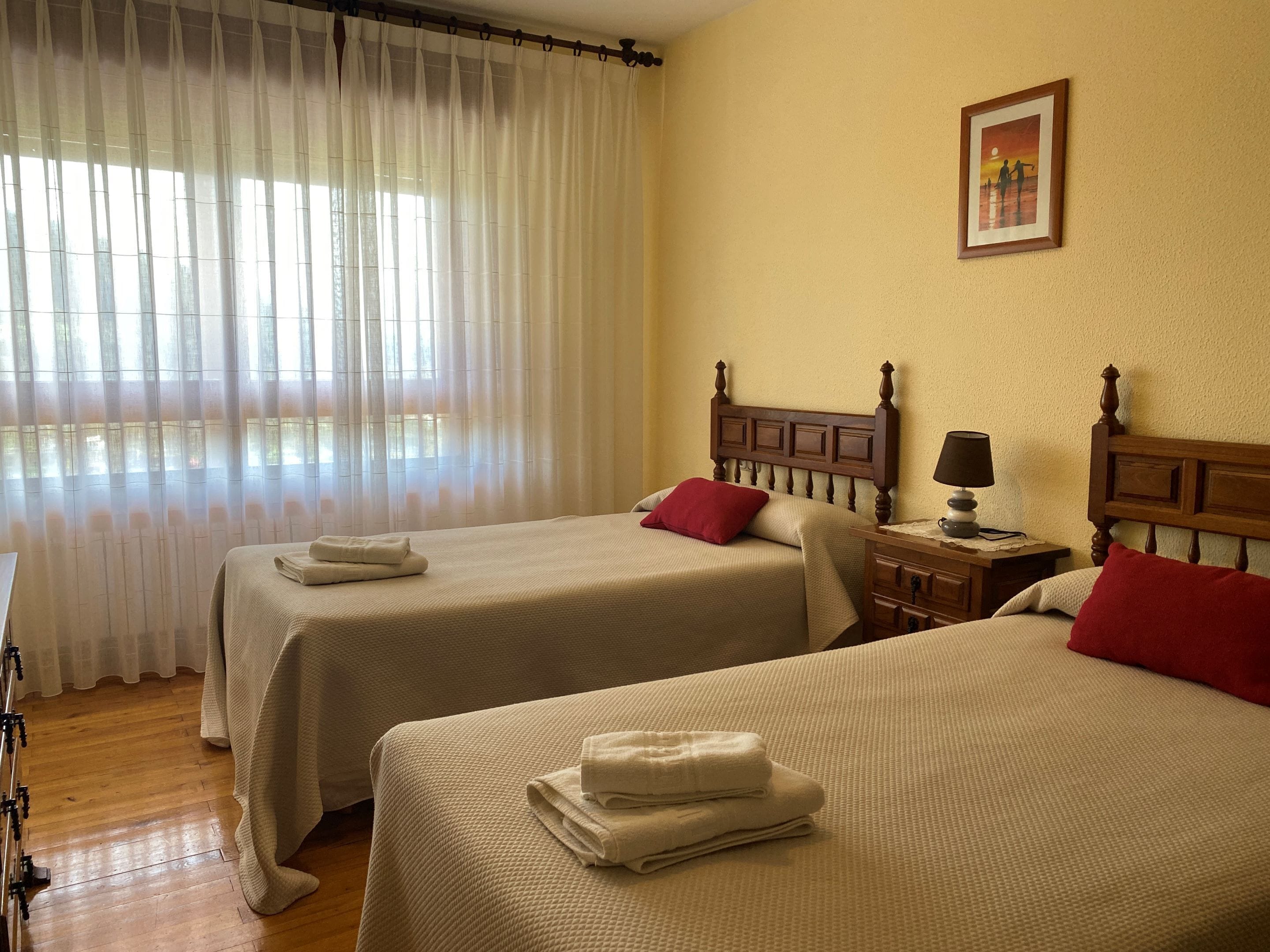 Quarto de hotel com duas camas, cabeceira de madeira, mesa de cabeceira com abajur e janela com cortinas translúcidas.