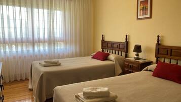 Quarto de hotel com duas camas, cabeceira de madeira, mesa de cabeceira com abajur e janela com cortinas translúcidas.