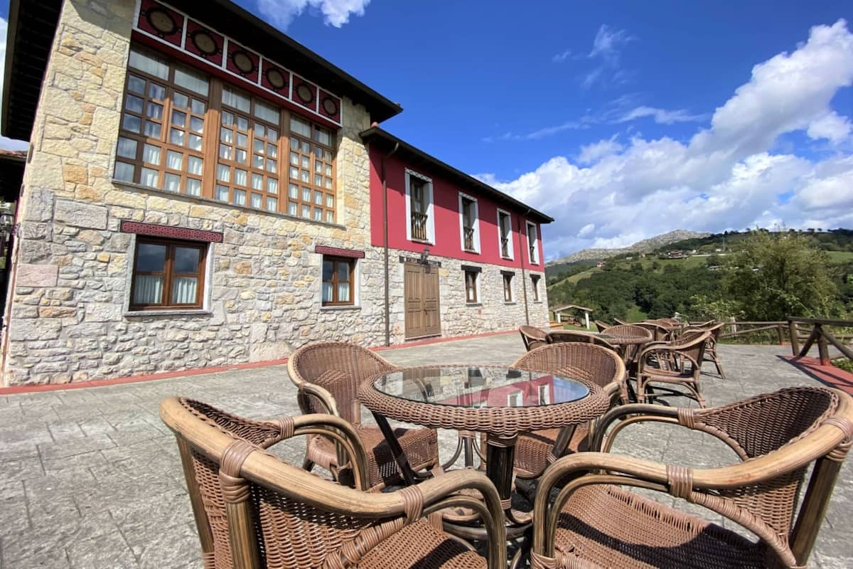 Un bâtiment en pierre avec une façade rouge et de nombreuses fenêtres. On y trouve une table ronde entourée de chaises en osier.