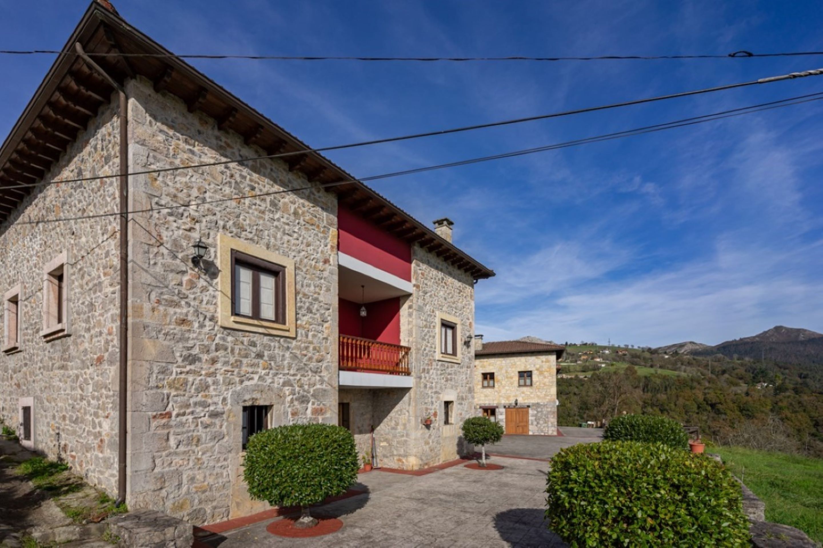 A stone building with a red balcony, surrounded by greenery and hills in the background.