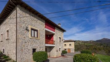 A stone building with a red balcony, surrounded by greenery and hills in the background.