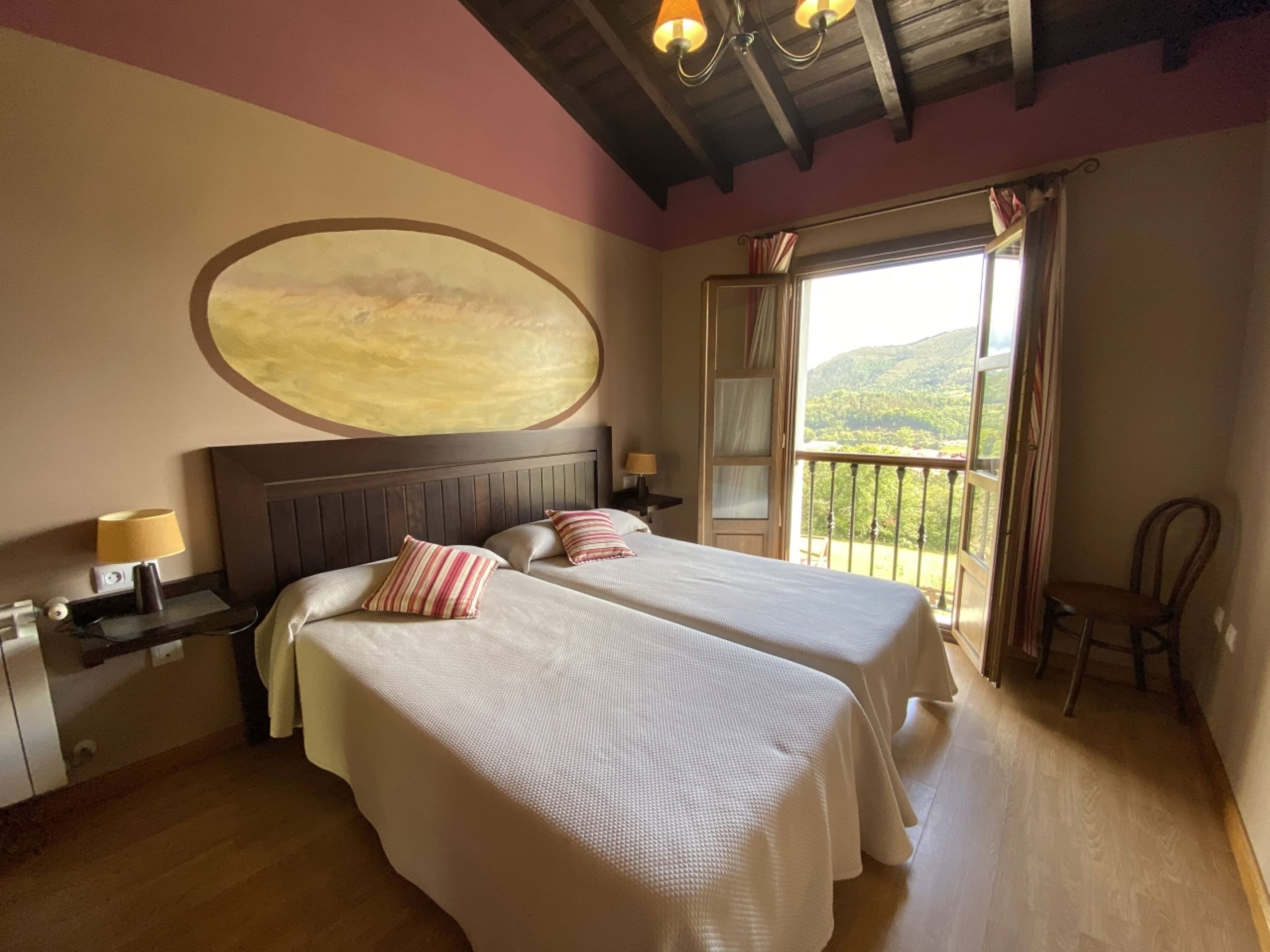 A bedroom with two beds, a large painting on the wall, a balcony with a view of the hills, and a wooden ceiling.