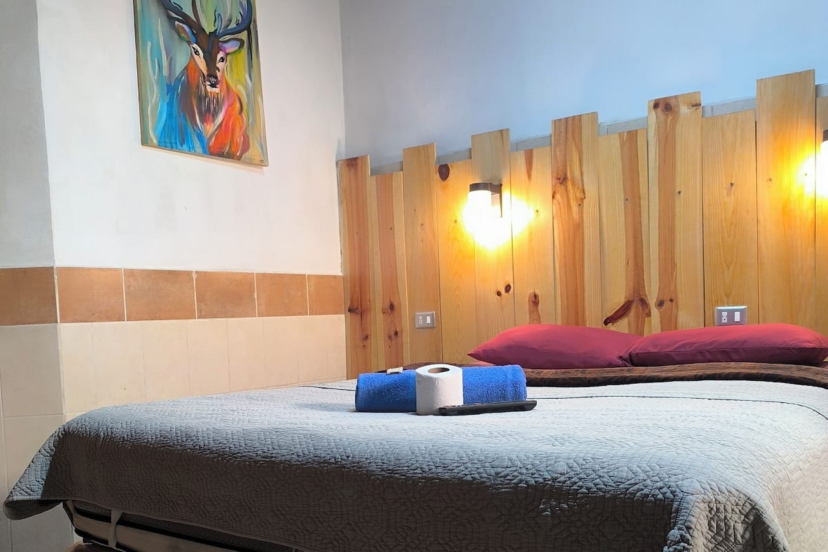 A hotel room with a bed, a wooden wall panel, a painting, and a wall-mounted light fixture.