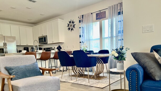 Un salon moderne avec un canapé gris, un fauteuil bleu et un canapé bleu orné de clous dorés. On aperçoit à l’arrière-plan un coin repas avec des chaises bleues et une table blanche.