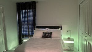 Une chambre à coucher comprenant un lit, une table de chevet, un ventilateur de plafond, une fenêtre avec des rideaux et une armoire.