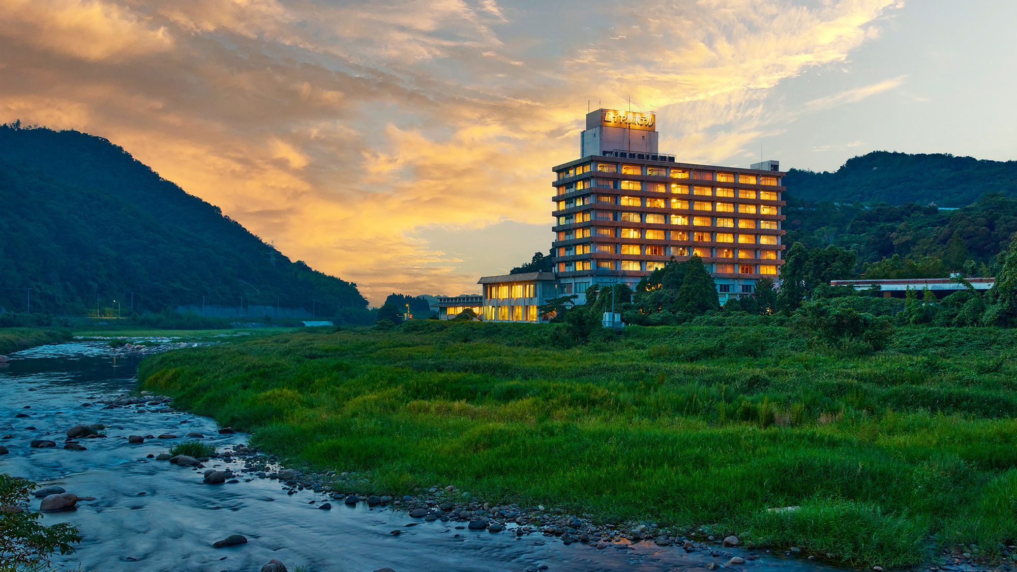 A multi-story hotel with a flat roof, surrounded by lush greenery and a river.