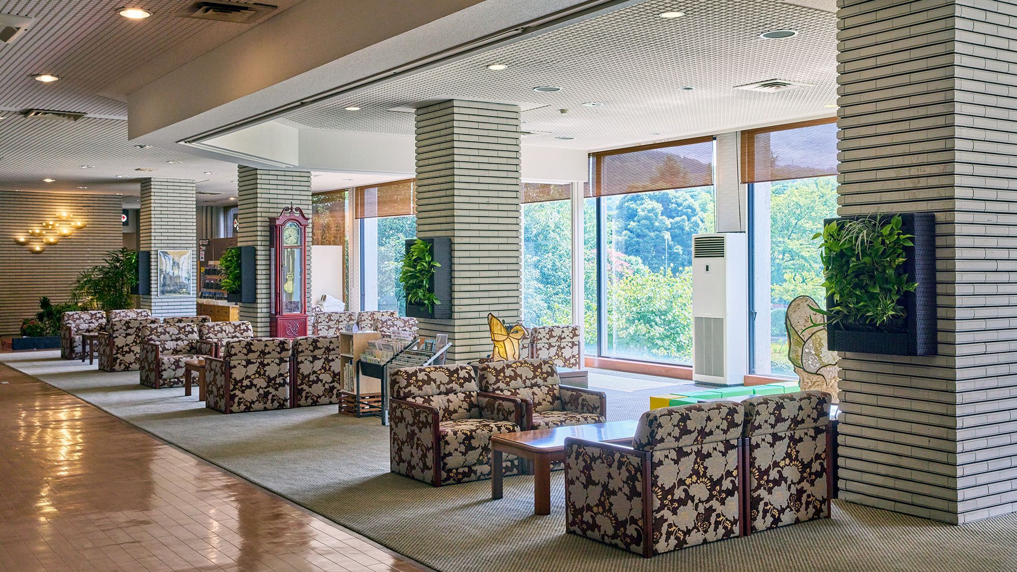 Een ruime lobby met een rij gestreepte banken en tafels, grote ramen met uitzicht op groen, en een modern plafondontwerp.