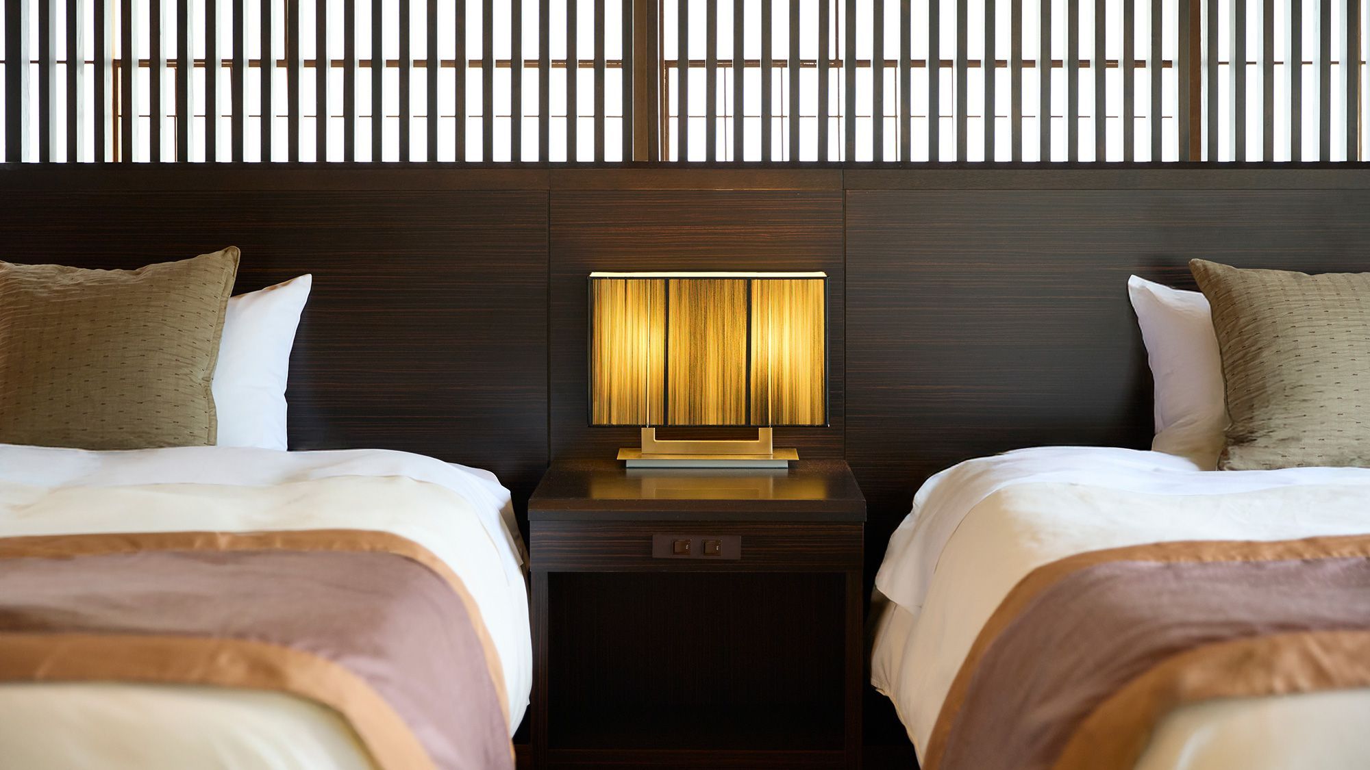 Habitación de hotel con dos camas, una mesita de noche con lámpara y cabecero de madera.