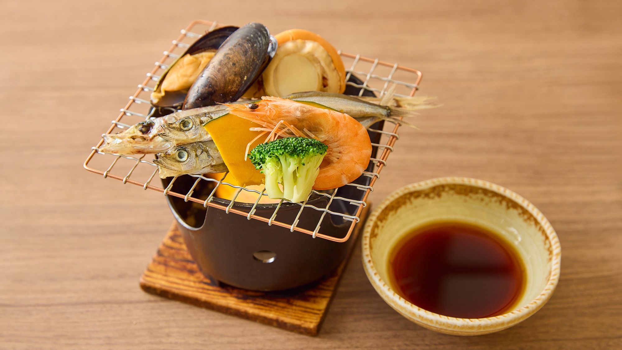 A variety of seafood on a wire rack, including shrimp, fish, and a clam, with a bowl of soy sauce beside it.