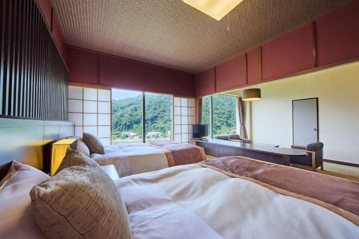 Habitación de hotel con dos camas, televisión, escritorio y ventanas grandes con vistas a zonas verdes.