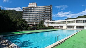 A swimming pool with a clear blue water surface, surrounded by a green lawn area. In the background, there is a multi-story building with a sign that reads 'ROYAL HOTEL'. - Misasa Royal Hotel (Misasa)