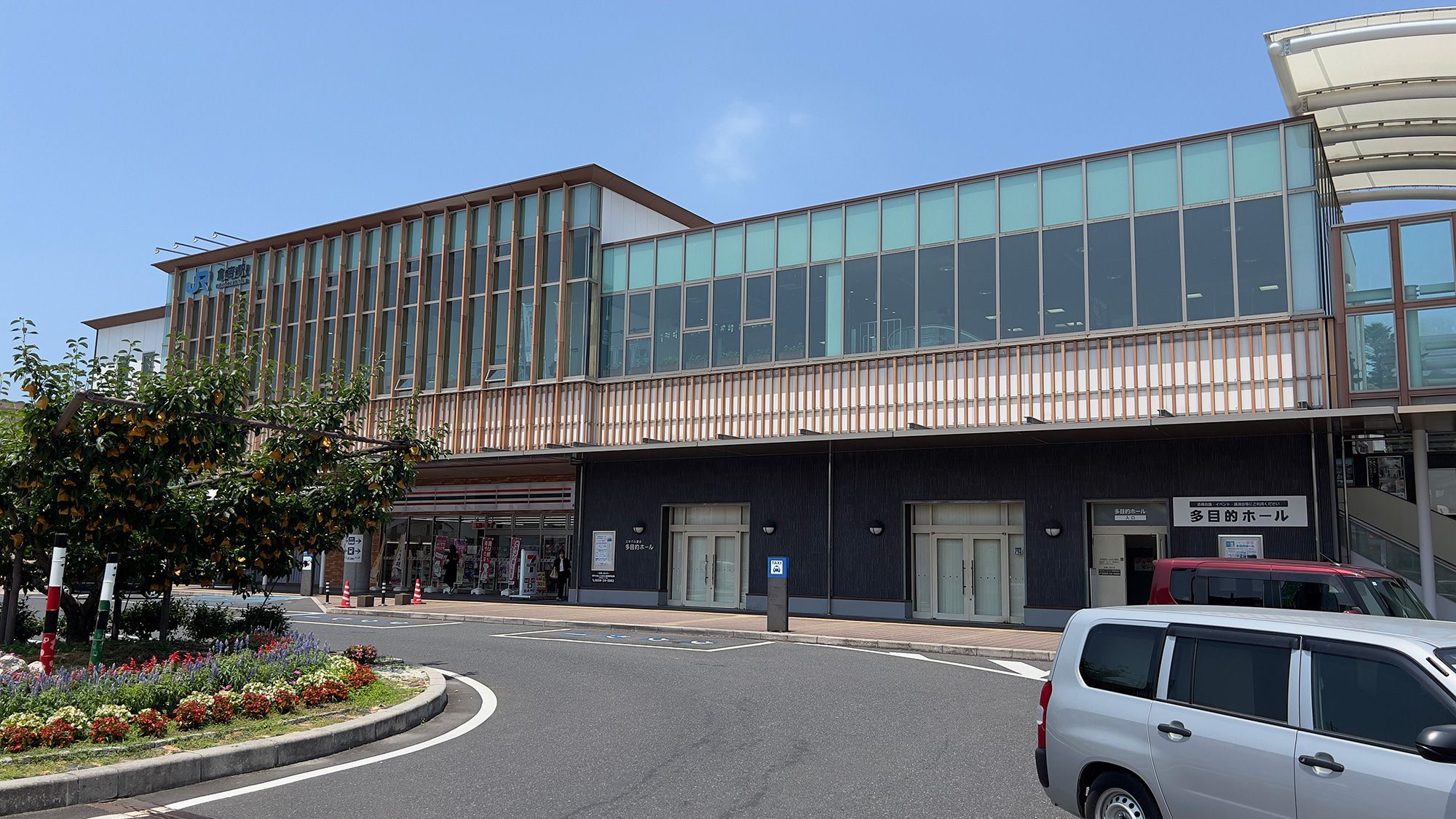 A modern building with a glass facade, a covered entrance, and a parking area with a white car.