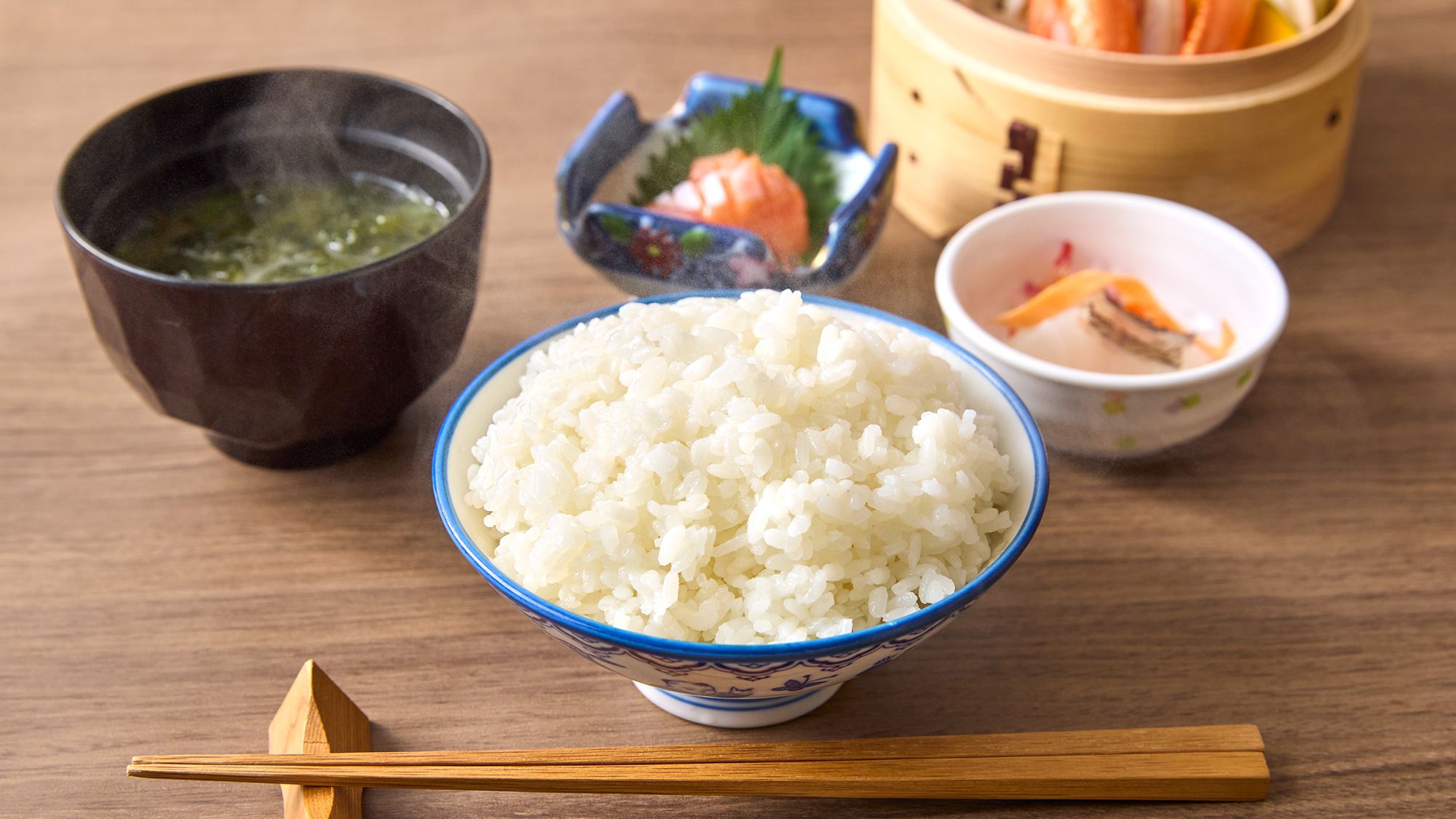 A bowl of white rice with chopsticks and a small bowl of soup on a wooden table.