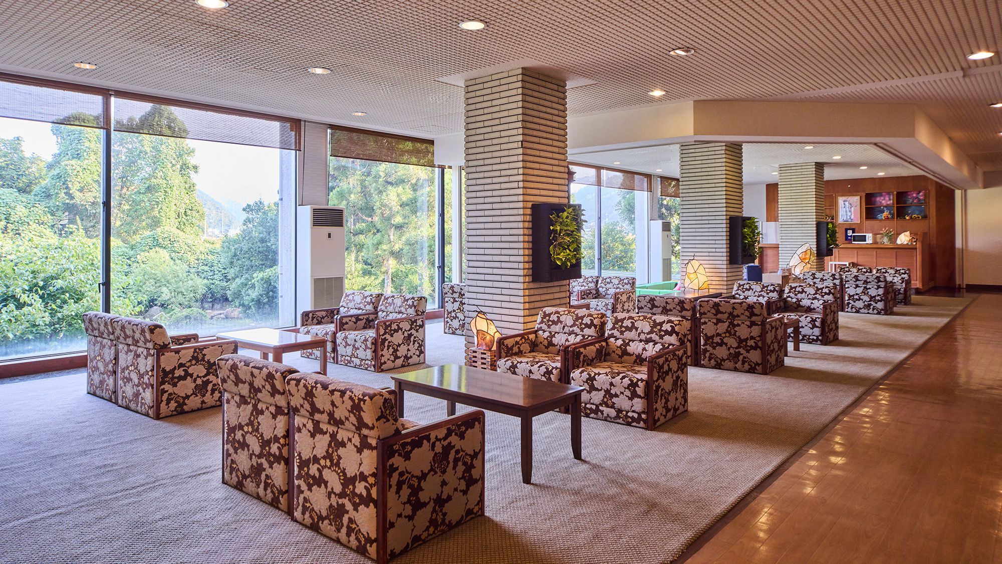 Un vestíbulo amplio con una larga fila de sofás y sillones, grandes ventanales que ofrecen vistas a la vegetación y un diseño de techo moderno.