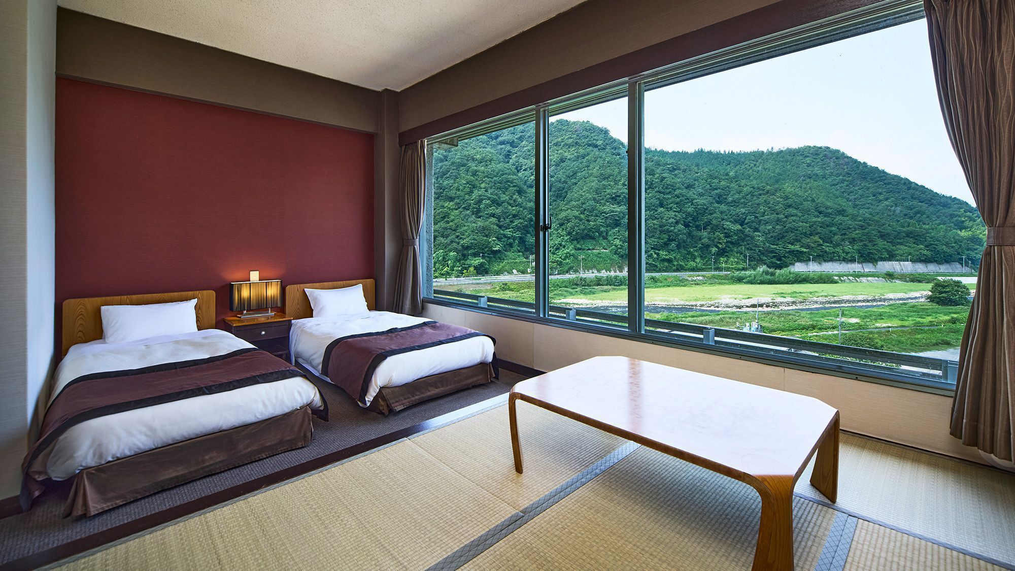A hotel room with two beds, a table, and a large window offering a view of a forested area.