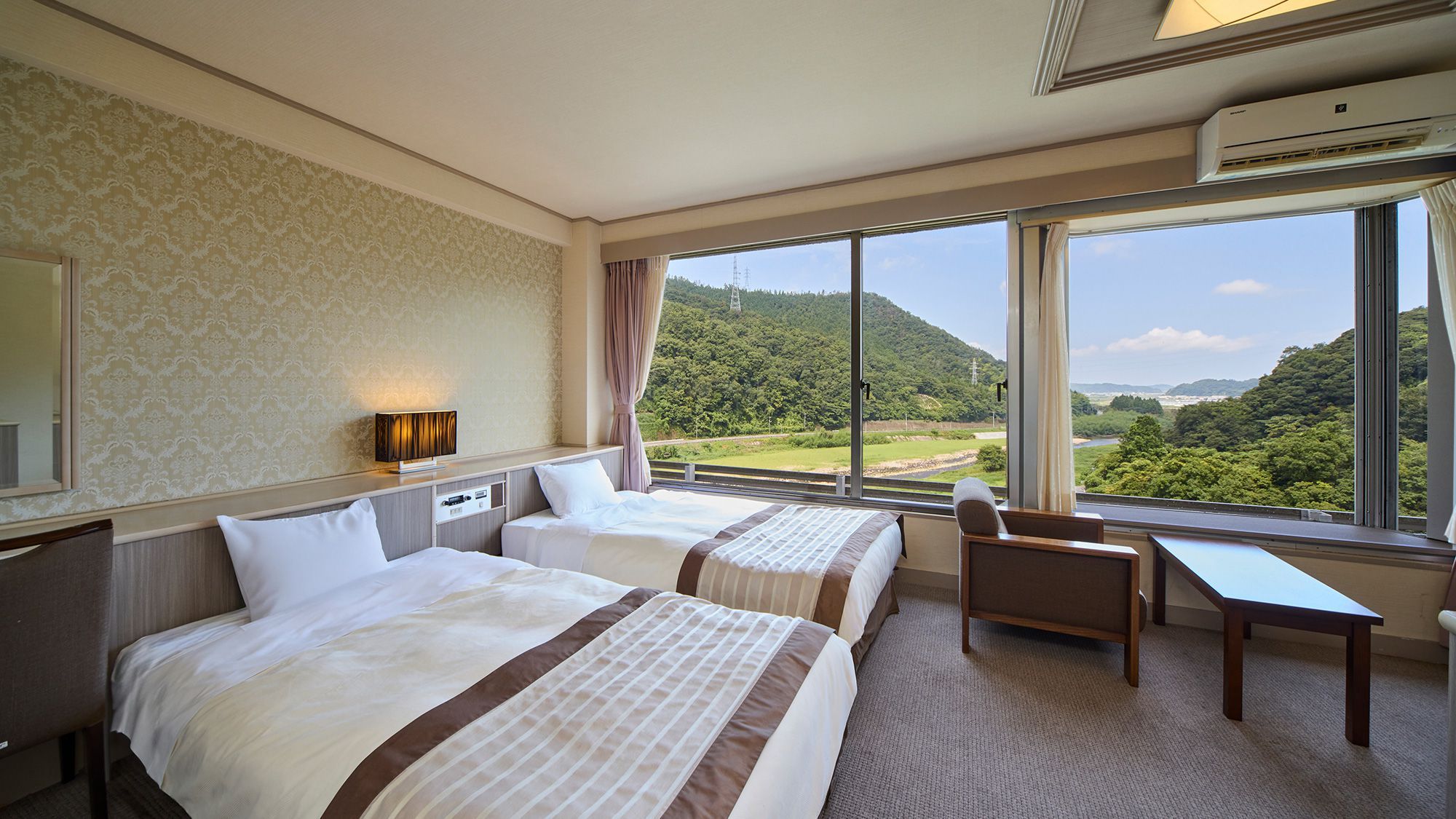 A hotel room with two beds, a desk, and a view of a green landscape.