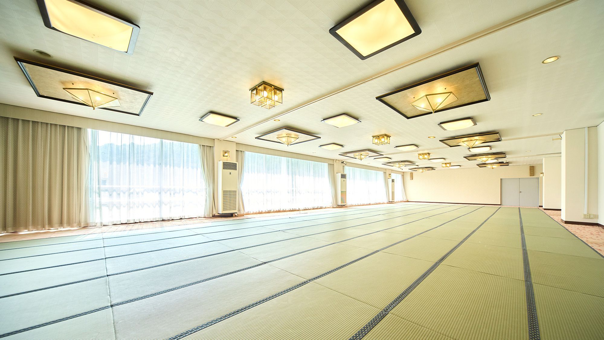 A spacious room with tatami mats, large windows with curtains, and recessed lighting.