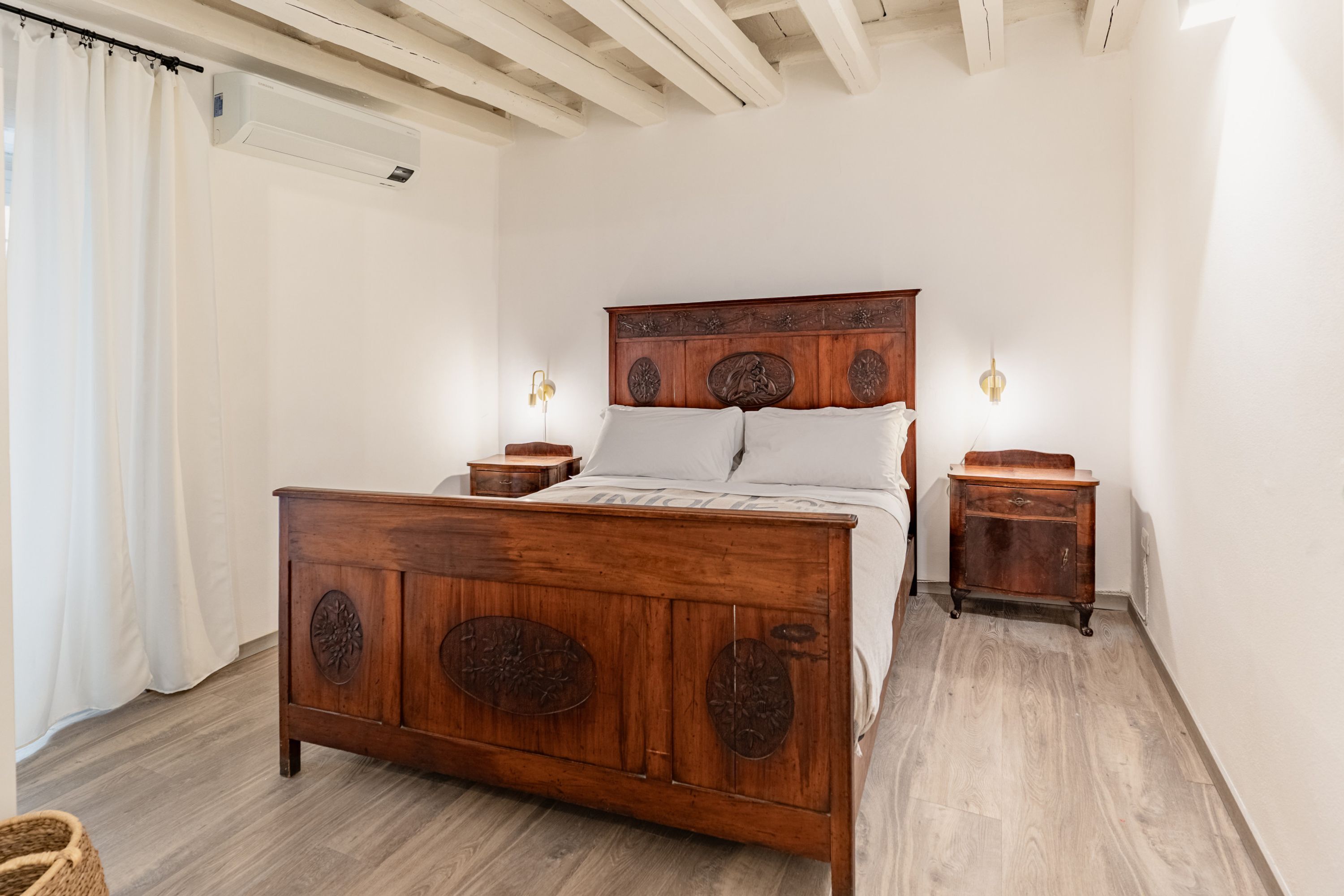 A bedroom with a wooden bed, white curtains, a small bedside table, and a wall-mounted light fixture.