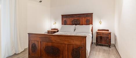 A bedroom with a wooden bed, white curtains, a small bedside table, and a wall-mounted light fixture.