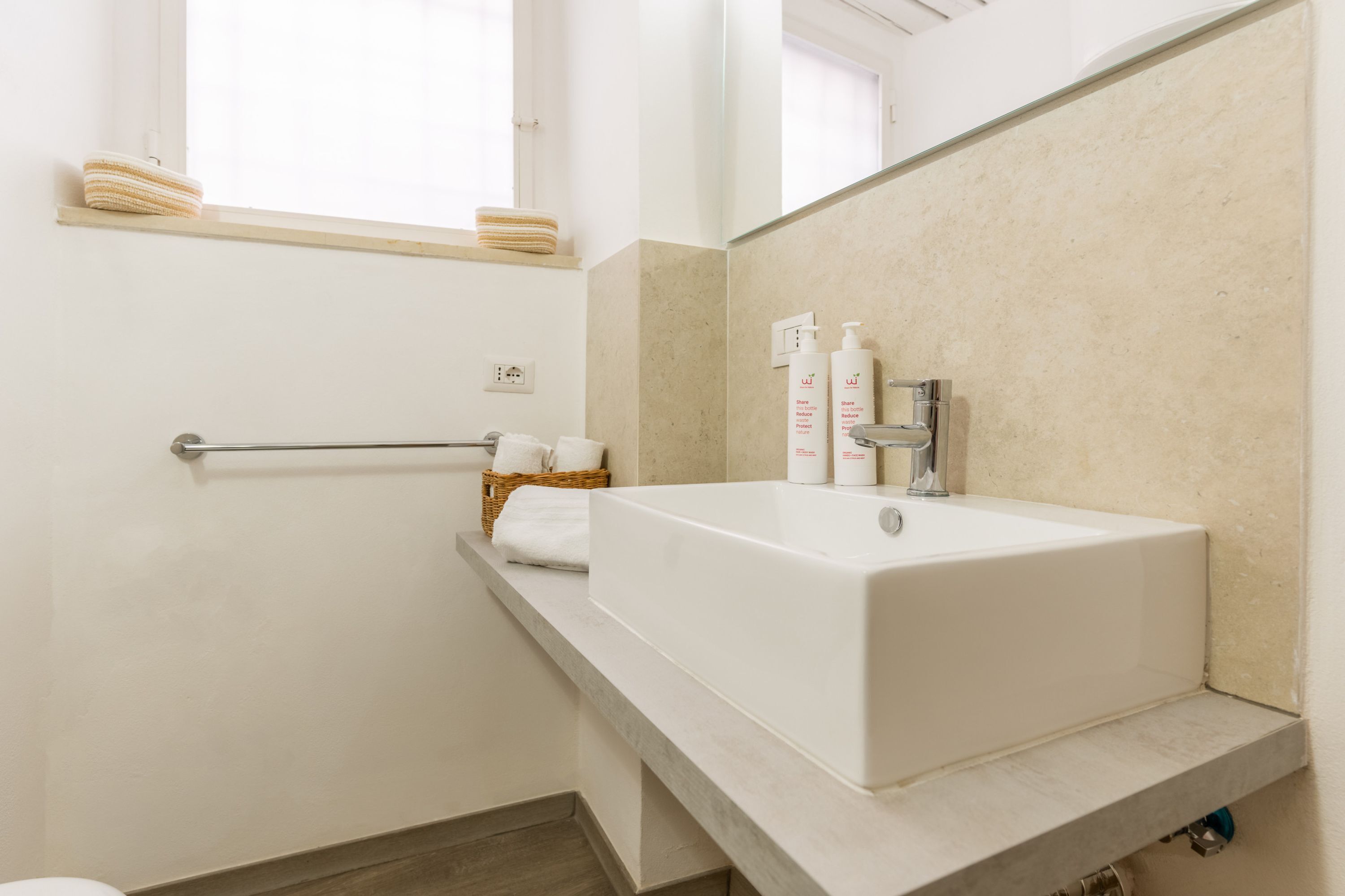 A modern bathroom with a white sink, beige tiles, a towel rack, and a window.