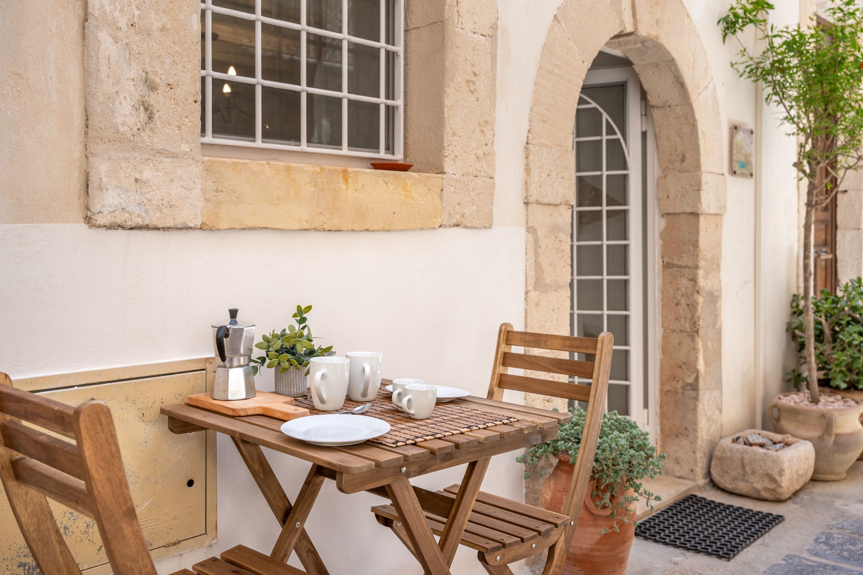 A wooden table set with plates, cups, and a coffee pot, positioned in a courtyard with stone walls and potted plants.