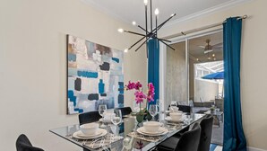A dining area with a glass table set for four, black chairs, a modern chandelier, and a large abstract painting on the wall. - Champions Gate Oasis 4BR Vacation Haven (Davenport)