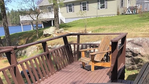 A wooden deck with a railing, two Adirondack chairs, and a view of a house and lake. - Rustic 2-bedroom cabin on Boyd Pond (Russell)