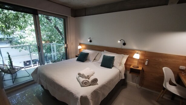 A neatly made bed with a rolled towel, two bedside lamps, and a view of the outdoors through a large window. - Estudio en Palermo III (Buenos Aires)