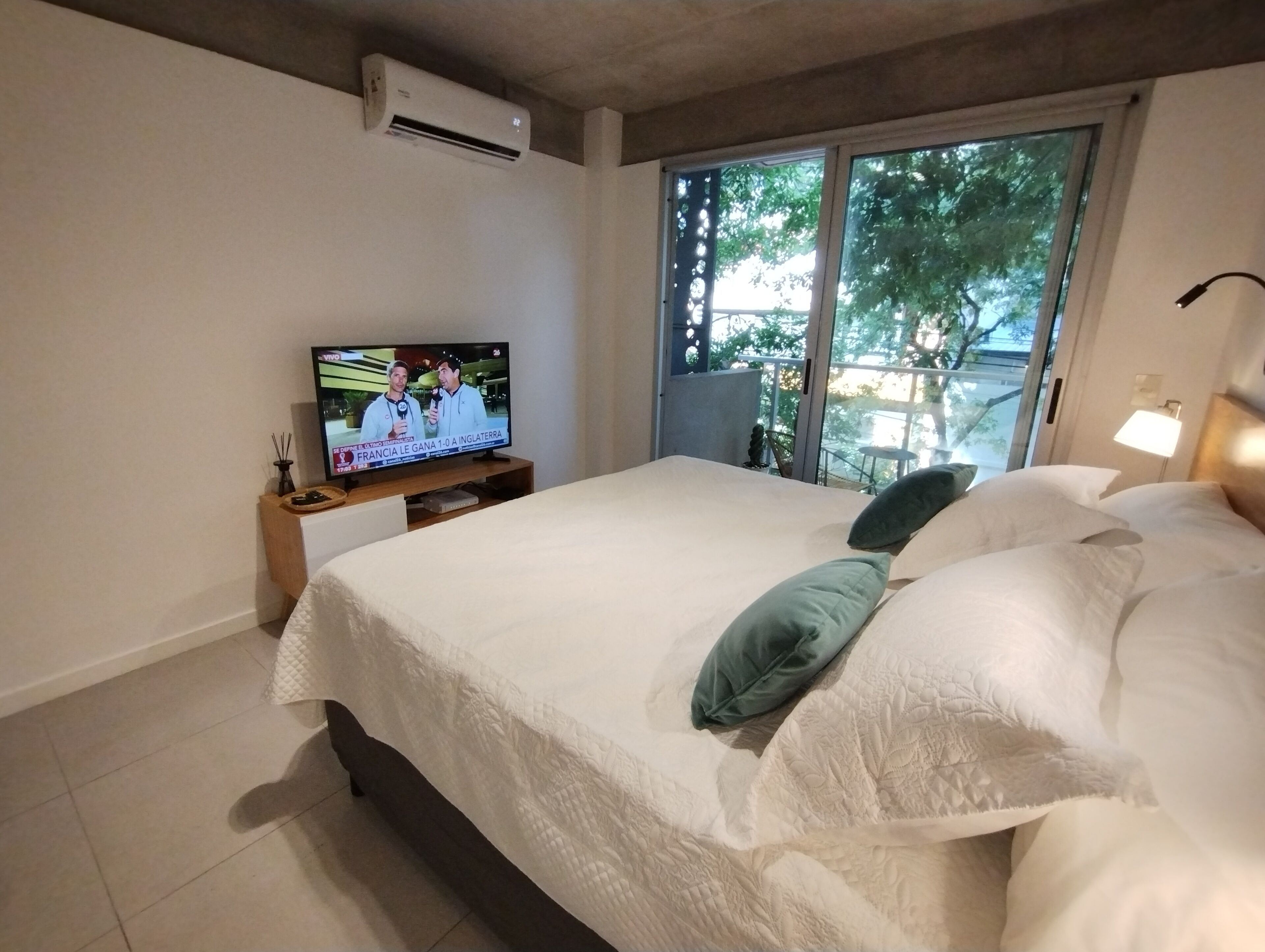 Ein Schlafzimmer mit Bett, einem Fernseher auf einem Holzgestell, einer Klimaanlage an der Wand und Blick ins Freie durch eine Schiebetür.