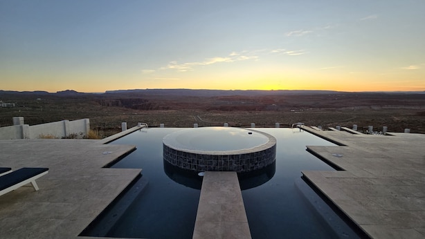 Une piscine à débordement avec un jacuzzi, entourée d’une terrasse et de bancs en béton, offrant une vue sur un paysage désertique au coucher du soleil.